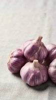 Fresh purple garlic bulbs with papery skin arranged on neutral linen fabric, suitable for food photography, culinary content, and cooking-related projects.