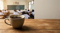 Coffee cup on wooden desk in blurred classroom with students and teacher at blackboard. Educational environment for morning break or teacher workspace concept.