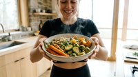 A young Caucasian woman with blonde hair, wearing a black t-shirt, smiles while holding a round woven basket filled with roasted vegetables including carrots, zucchini, and herbs in a bright kitchen.