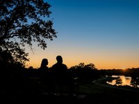 Silhouetted couple sitting together on bench overlooking river at sunset, watching golden sky reflect on water during romantic evening outdoors.