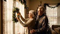 Senior Caucasian couple decorating home with Christmas wreath by window, embracing during holiday preparation in cozy living room with festive garland decorations.