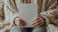 Woman in cozy knit sweater holding blank white book while sitting. Manicured hands visible against soft beige cable knit texture. Minimalist lifestyle reading moment.
