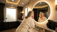 Senior woman in bathrobe applying skincare routine at illuminated vanity mirror in luxury bathroom spa setting for wellness and self-care concept.