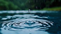 Water drop falling into calm blue water surface creating concentric ripples and splash crown against blurred dark green natural background.