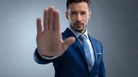Confident Caucasian businessman in blue suit showing stop gesture with palm, expressing authority and boundary setting in professional environment.