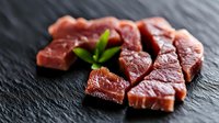Raw fresh beef chunks arranged on black stone surface with green herb garnish, showing meat texture and marbling. Culinary and food photography concept.