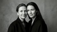 Two Caucasian women, senior mother and adult daughter, pose together in black outfits against a neutral backdrop in a timeless black and white portrait.