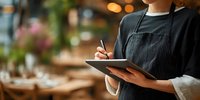 Female server in black apron taking order on tablet in restaurant with warm ambiance and blurred dining tables in background.