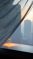 Glowing candle on windowsill at sunset with sheer curtain and city silhouette in background creates peaceful atmospheric mood for wellness and meditation concepts.