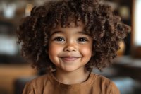 Happy mixed-race toddler girl with curly afro hair smiling at camera in warm brown sweater, portrait shot with soft natural lighting for family content.