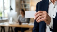 Mature male professional in business attire holding blank business card in modern office environment with colleagues working in background, suitable for corporate networking and branding.