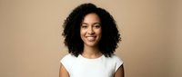 Young African American woman with curly hair smiling confidently in white top against beige background for professional headshot and business portrait.