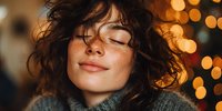 Young woman with curly hair enjoying peaceful moment with warm bokeh lights in background. Cozy winter portrait for lifestyle and wellness content.
