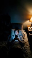 Young man using smartphone in bed at night with warm lamp light creating cozy bedroom atmosphere for sleep hygiene and technology addiction concepts.