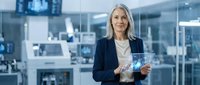 Middle-aged woman with gray hair, Caucasian ethnicity, wearing dark blazer and white blouse, holding digital tablet in modern office environment with glass partitions and cityscape view.