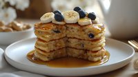Stacked pancakes topped with blueberries and banana slices, drizzled with maple syrup on a white plate for breakfast and brunch dining.