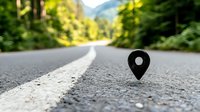 Location pin marker on empty mountain road with blurred forest background, low angle view focusing on black navigation symbol on asphalt surface with white line marking.