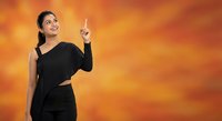 Young Asian businesswoman in black dress pointing upward with finger against orange gradient background for presentations and marketing materials.
