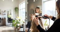 Professional hairstylist styling blonde woman's hair with curling iron in modern bright salon interior with natural lighting and plants.