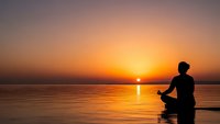 Silhouette of person meditating in lotus position by calm water during golden sunset. Peaceful mindfulness and wellness concept for relaxation practices.