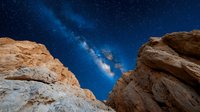Dramatic night sky view of Milky Way galaxy between red rock formations in desert landscape, stars and cosmic clouds illuminating dark blue space.