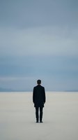 Businessman in dark suit standing alone on white sand beach under cloudy sky, rear view solitude concept for business motivation and leadership themes.