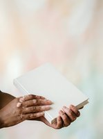 Hands of adult male with medium skin tone holding open white book against soft blurred background, suitable for education, learning, reading, and literacy concepts.
