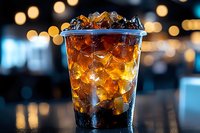 Cola drink with ice cubes in clear plastic cup, served in modern cafe or restaurant setting with blurred bokeh lights in background.