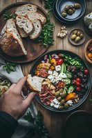 Fresh Mediterranean bowl with artisanal bread, feta cheese, olives, colorful vegetables, and cold cuts arranged on rustic wooden table. Hands reaching for crusty bread.