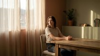 Young woman sitting at wooden table by window with warm sunlight streaming through sheer curtains in cozy home interior workspace.