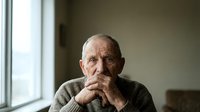 Thoughtful elderly Caucasian man with hands clasped under chin, sitting by window in contemplative pose. Senior male portrait for healthcare concepts.