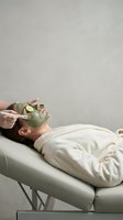 Young Caucasian woman lying on spa table in robe receiving green clay facial mask with cucumber slices, skincare beauty treatment.