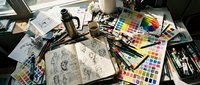 Artist workspace with open sketchbook featuring botanical drawings, surrounded by watercolor palettes, brushes, pencils, and design materials for creative projects and illustration work.