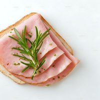 Open sandwich with sliced ham and fresh rosemary on white bread against white background, garnished with herbs for breakfast or lunch.