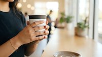 Young woman with medium skin tone holding a reusable coffee cup with black lid in bright kitchen space, suitable for lifestyle, sustainability, and beverage marketing content.
