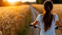 Young woman in white t-shirt riding bicycle through golden wheat field at sunset, view from behind capturing rural summer atmosphere and healthy lifestyle.