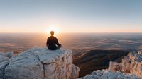 Man sitting on rocky cliff edge meditating during golden hour sunrise overlooking vast mountain valley landscape for wellness and mindfulness content.