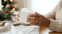 Hands holding white gift box with ribbon during holiday season celebration with decorated tree and warm lights in background.