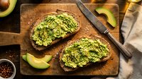 Avocado toast on dark bread with mashed avocado spread, healthy breakfast meal on wooden cutting board with knife and fresh avocado slices.