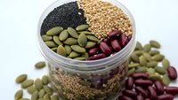 Glass jar filled with variety of healthy seeds and legumes including black sesame, quinoa, pumpkin seeds and red kidney beans for cooking.