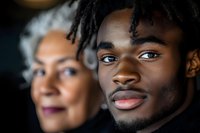 Young African American man with dreadlocks in foreground, mature woman blurred behind, warm lighting creates intimate family portrait atmosphere.