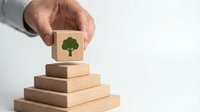 Hand placing wooden block with green tree icon on pyramid of blocks representing environmental protection and sustainable growth concept.