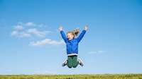 Young child with blonde hair, Caucasian ethnicity, wearing blue jacket and green pants, jumping with arms raised in celebration outdoors on grass field under clear sky.