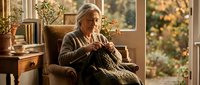 Senior woman with gray hair knitting in armchair by window with autumn garden view, cozy home interior with plants and natural lighting.