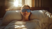 Young woman wearing blue sunglasses lying in bed with white bedding, illuminated by warm morning sunlight streaming through window creating dramatic lens flare effect.