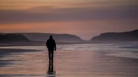 Surfer in wetsuit standing on wet sand beach at sunset, holding surfboard, coastal landscape with cliffs and ocean waves.