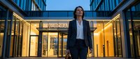 Professional woman in business attire walking confidently through modern glass corridor with illuminated storefront at dusk, suitable for corporate and entrepreneurship concepts.