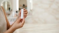 Woman holding white lip balm stick in bathroom setting with mirror and candles in background for skincare and beauty product promotion.