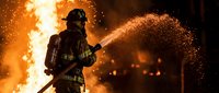 Firefighter in protective gear spraying water on massive flames during emergency rescue operation, intense heat and sparks flying.
