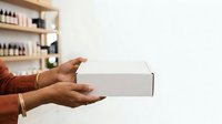 Person with medium skin tone holding white product box against neutral background with shelves of bottles, suitable for e-commerce, retail, packaging, and product marketing campaigns.